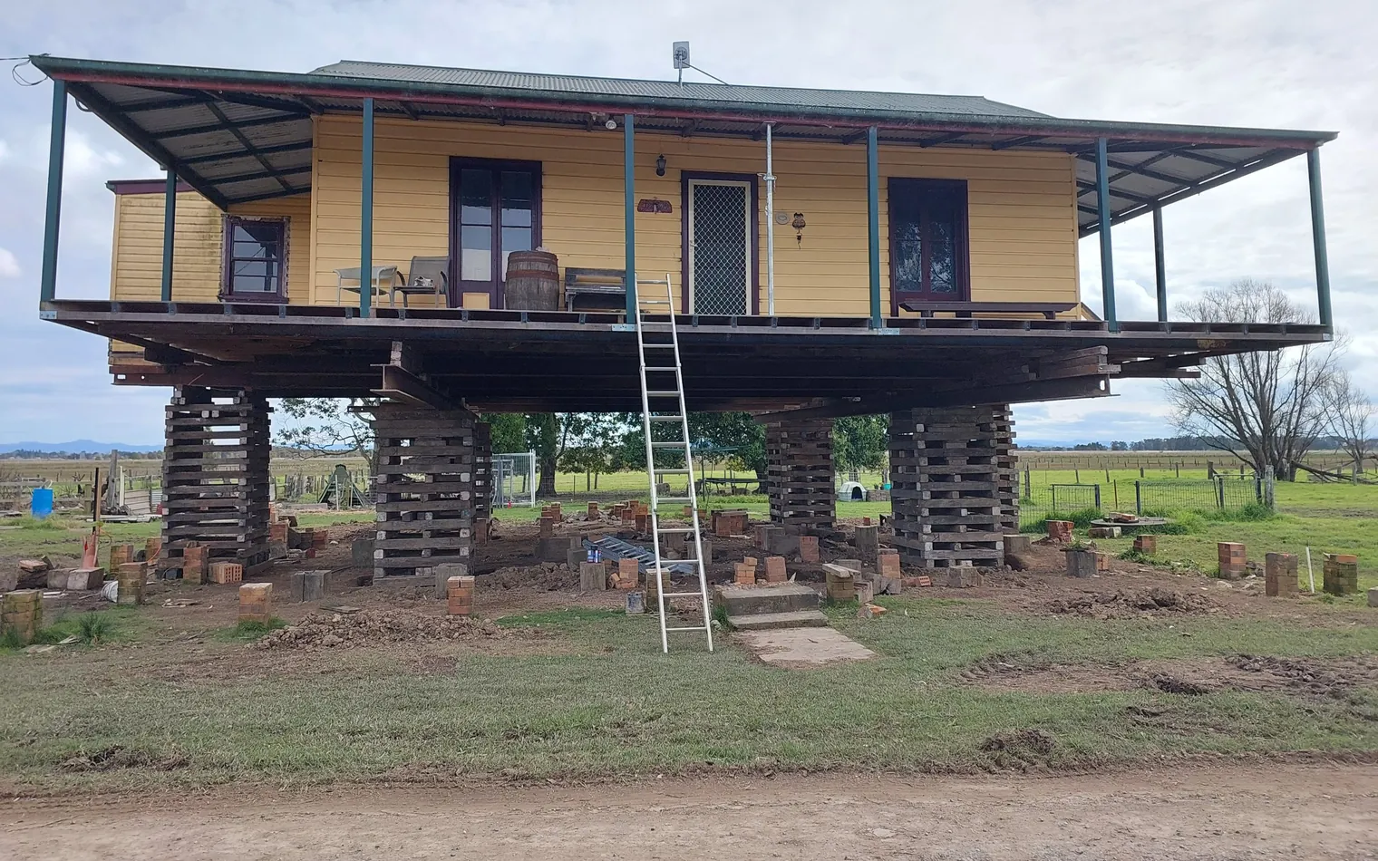 Yellow weatherboard farmhouse with wraparound verandah raised on timber cribbing during a house lift